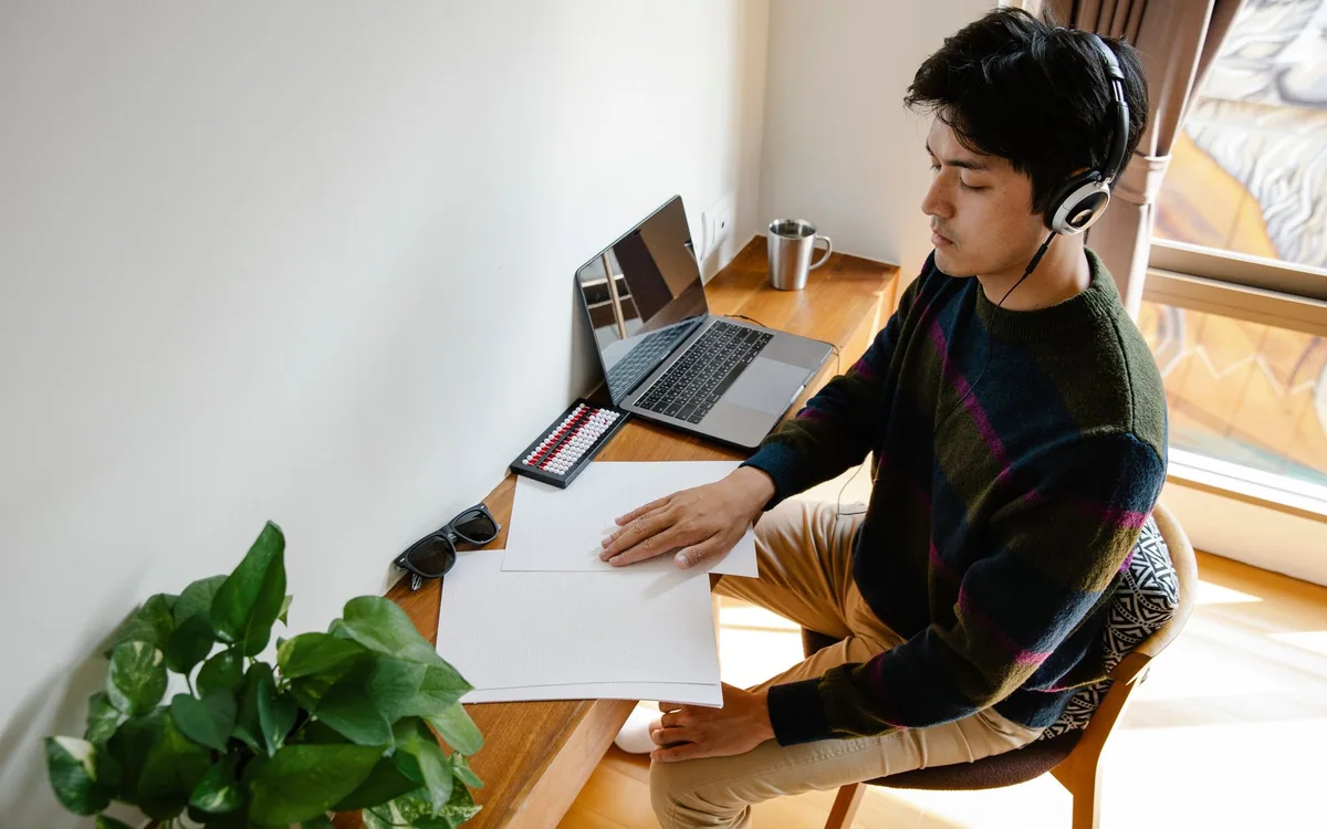 A man wearing over-ear headphones works at a desk beside plants.