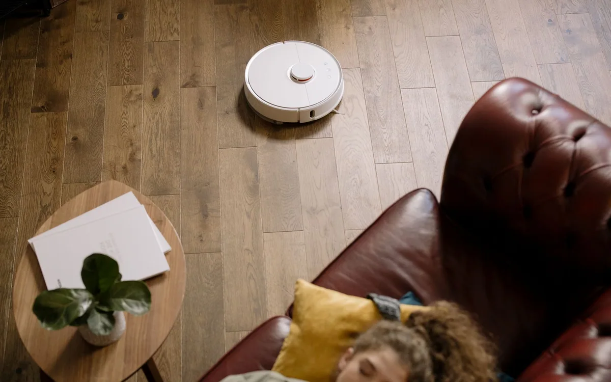 A robot vacuum on a wooden living room floor while a woman rests on a sofa.
