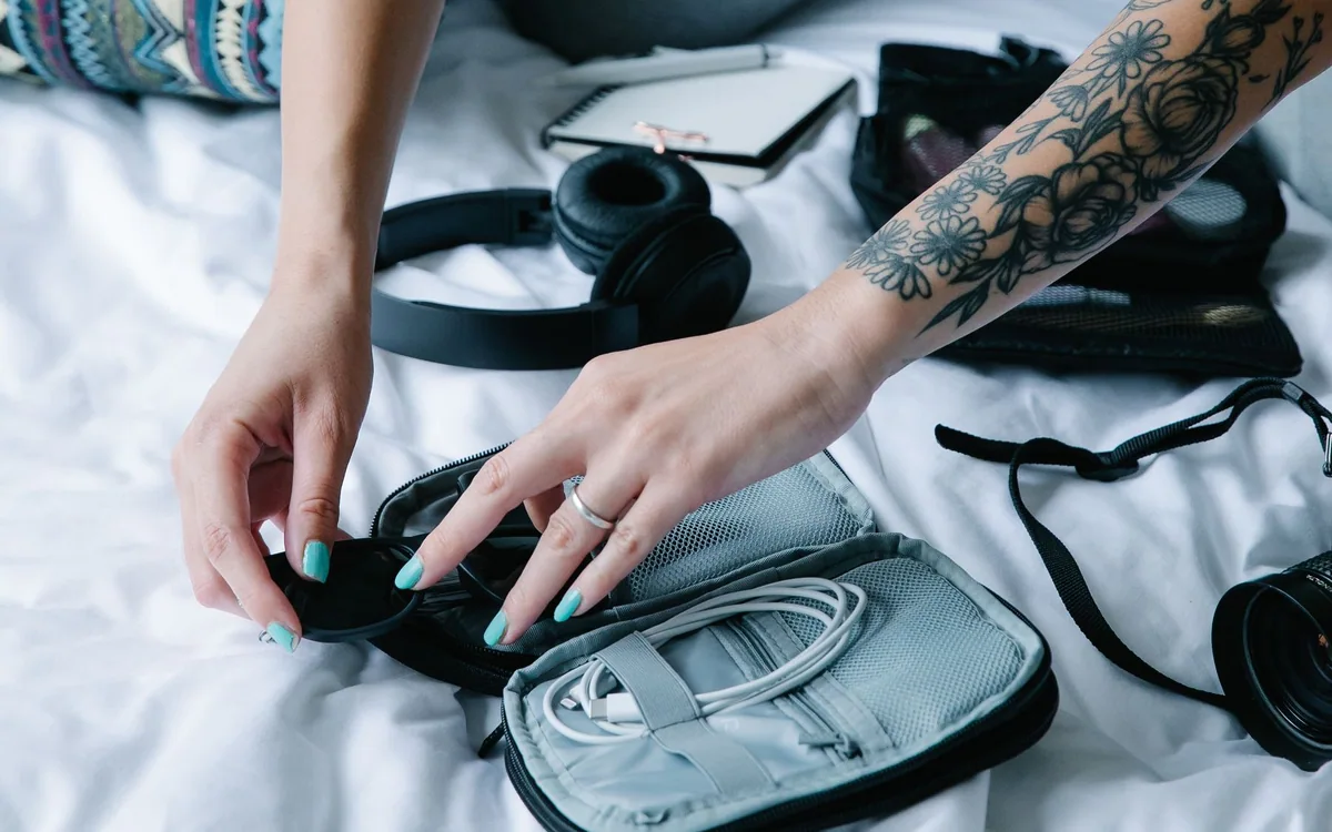 Hands organize a gray electronics pouch and accessories on a bed.