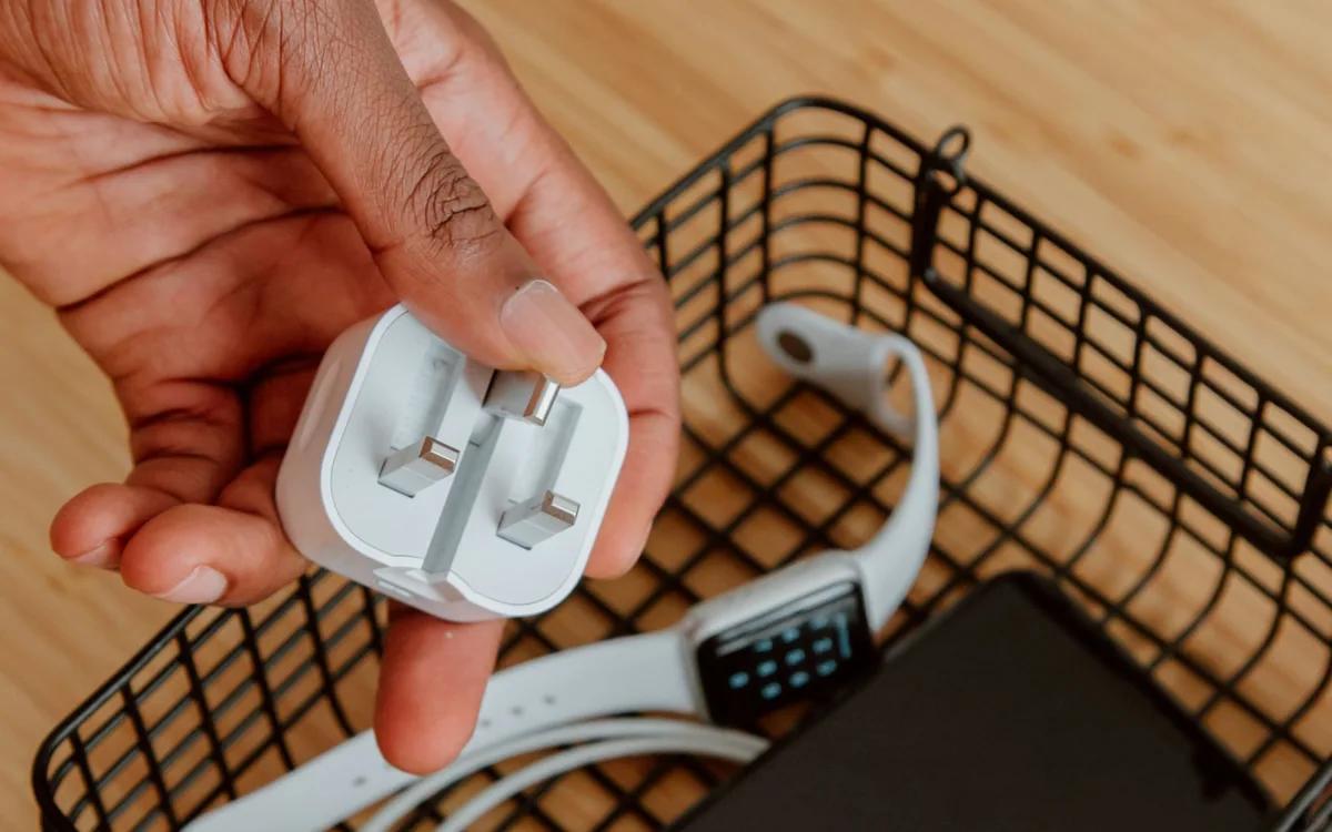 A hand holds a charging plug beside a basket containing a smartphone and watch.